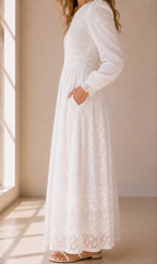 A woman wearing a long-sleeve white lace maxi dress with side pockets, standing in a sunlit room with beige walls and floor.