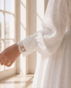 A close-up of a person wearing a white textured long-sleeve dress or robe with delicate lace trim at the cuff, standing near a sunlit window.