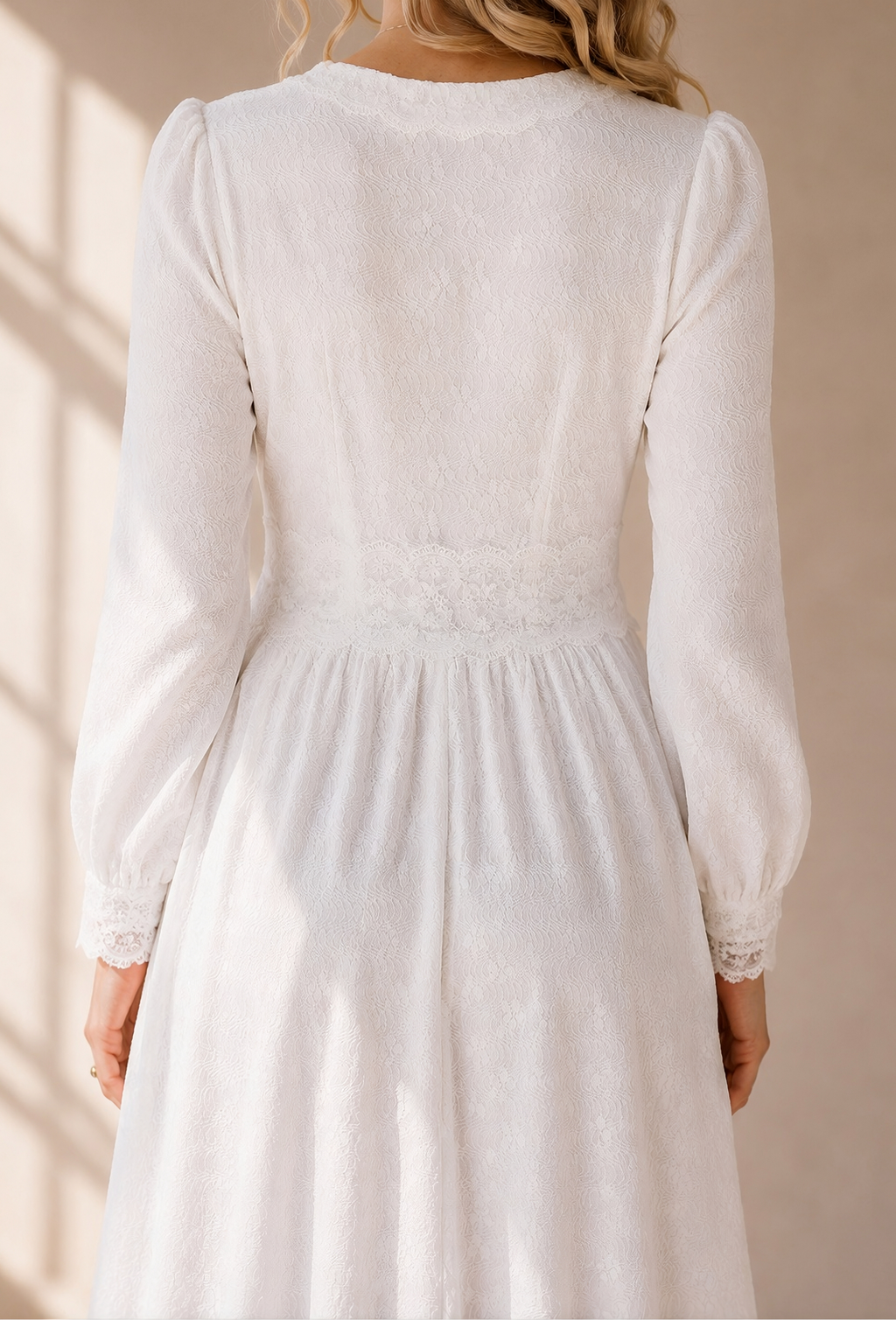 A woman wearing a white long-sleeve midi dress with a gathered waist and lace trim, shown from the back in warm sunlight against a beige wall.