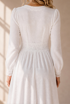 A woman wearing a white long-sleeve midi dress with a gathered waist and lace trim, shown from the back in warm sunlight against a beige wall.