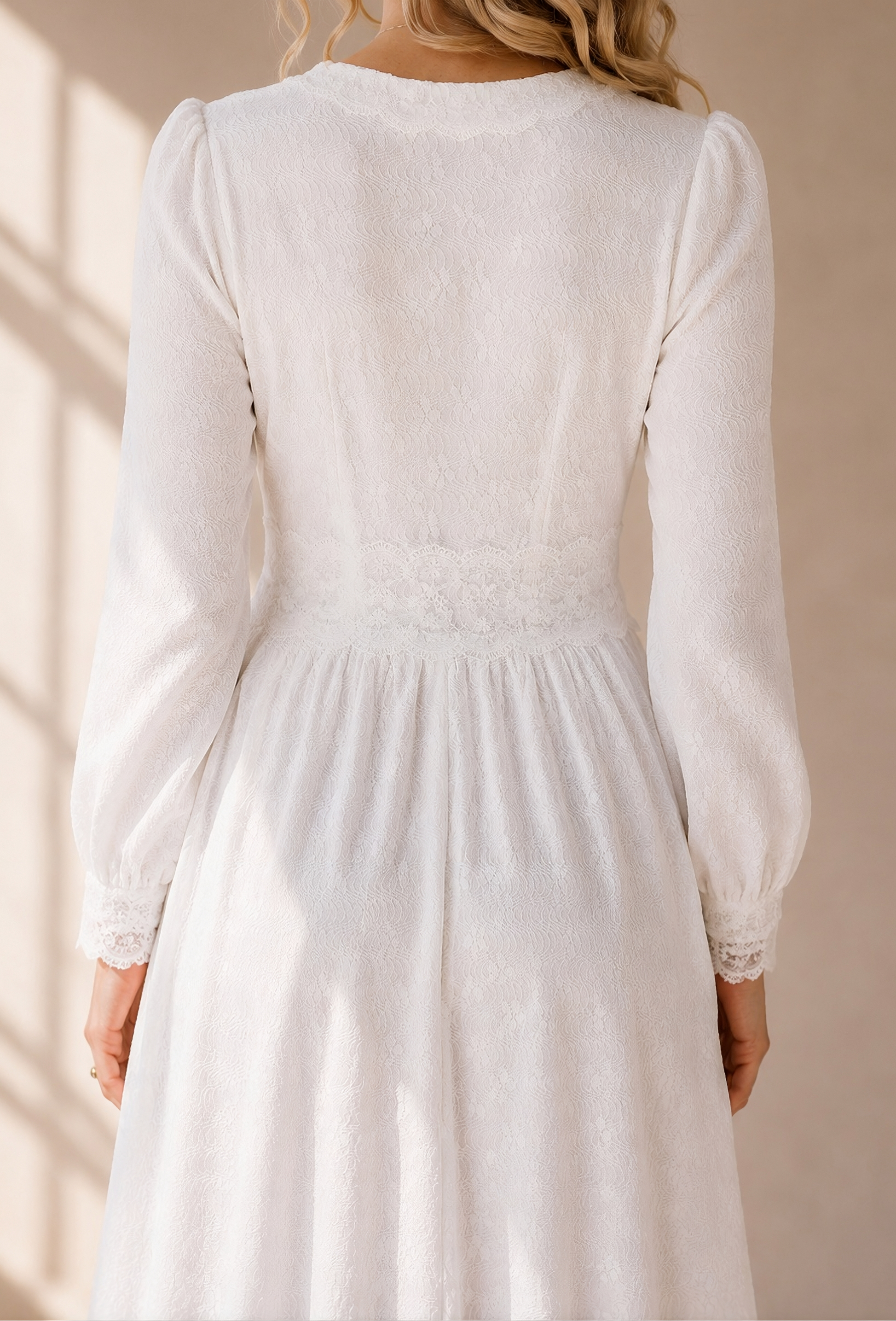 A woman wearing a white long-sleeve midi dress with a gathered waist and lace trim, shown from the back in warm sunlight against a beige wall.